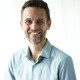 Smiling person with short curly hair and beard, wearing a light blue button-up shirt, against a white background.