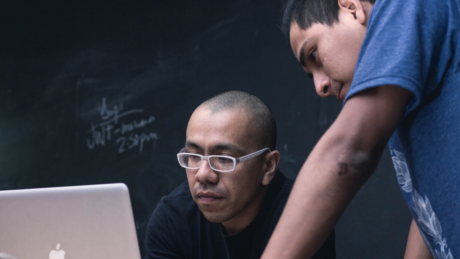 Two men look at a laptop in front of a blackboard.