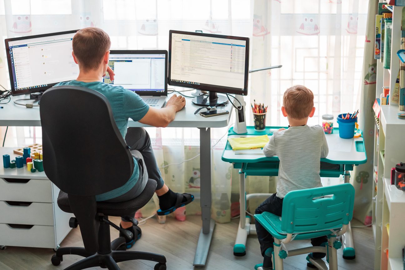 A stay-at-home dad works at his office desk while his son does homework next to him.