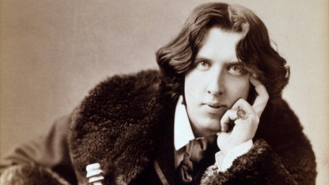 A man with a fur coat is posing thoughtfully with his hand on his chin.