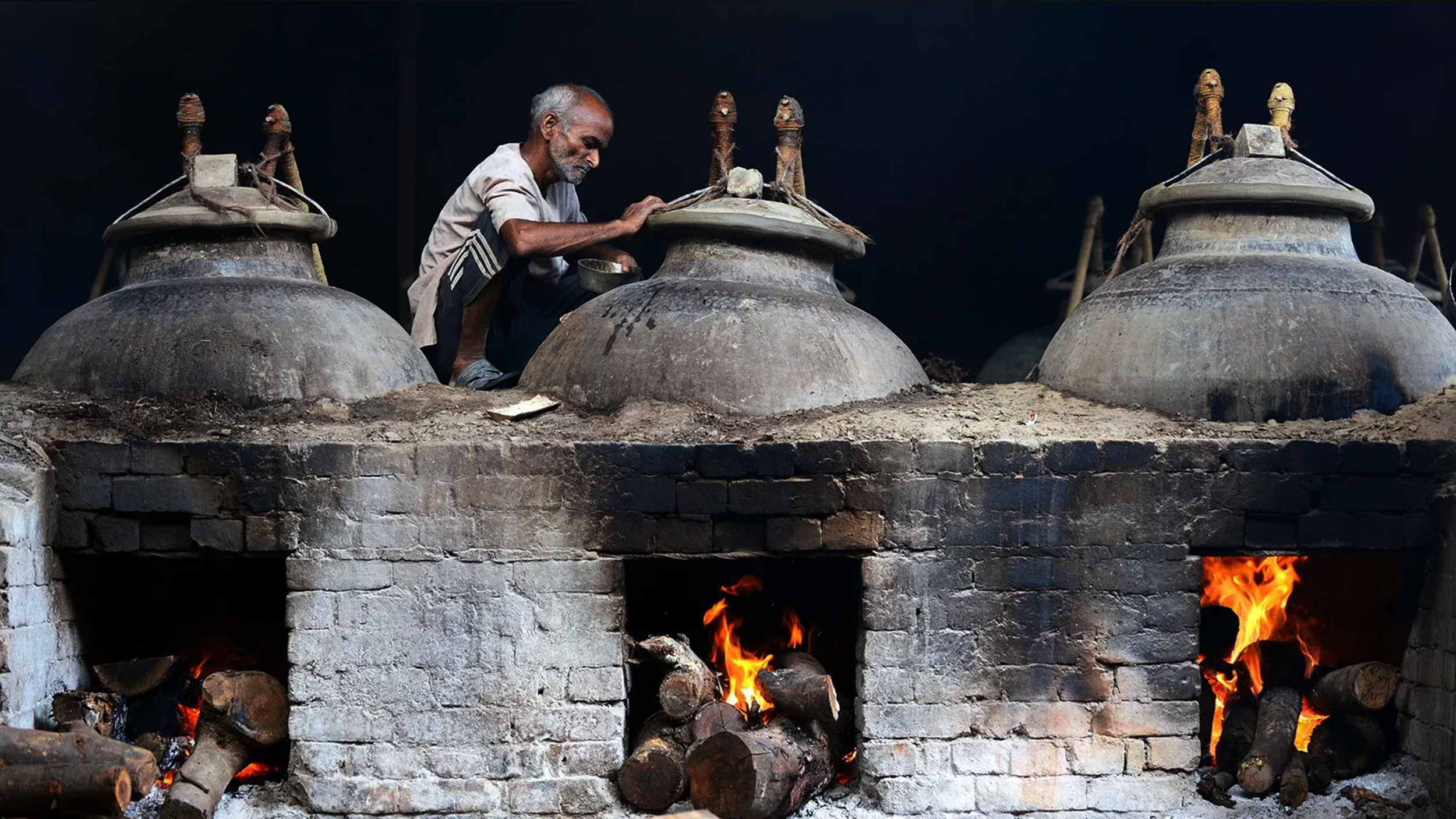 A man is using mitti attar on a clay pot in front of a fire.
