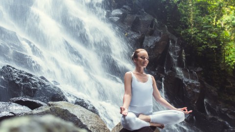 A woman peacefully meditating beside a gushing waterfall.