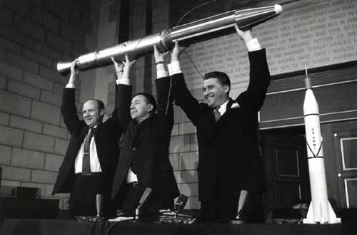 Three men holding up a rocket in front of a table.