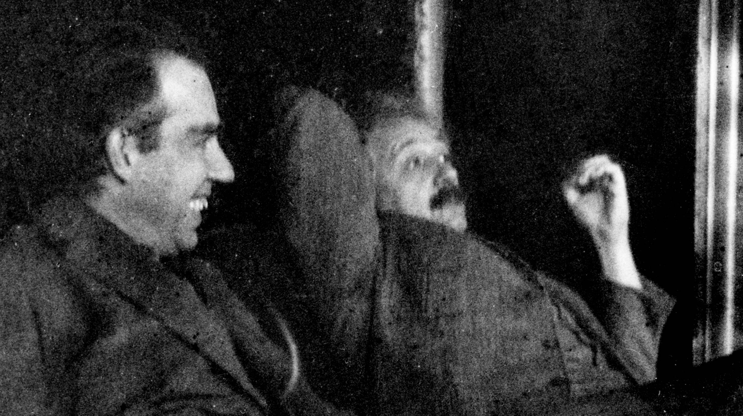 Two men sit closely together, one smiling and the other reclining with a relaxed posture against a dark background.