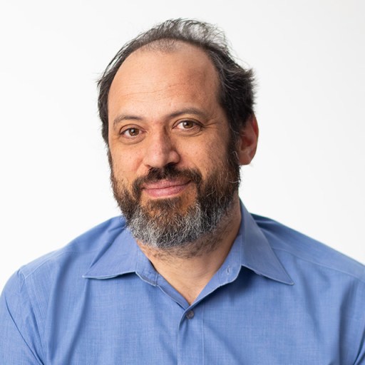 A man with a beard and a receding hairline wearing a light blue collared shirt, looking at the camera against a white background.