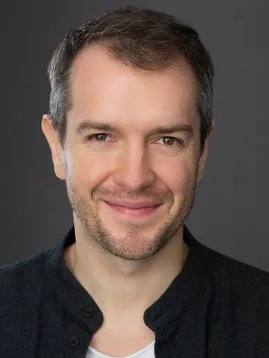 A headshot of a smiling man with stubble wearing a dark-colored shirt over a white t-shirt against a gray background.