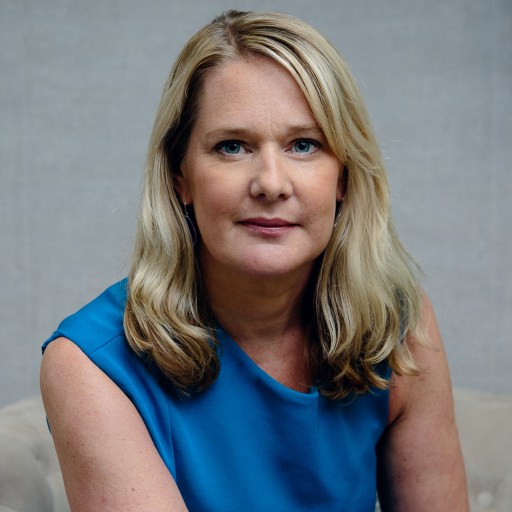 A woman with blond hair wearing a blue top poses for a portrait in front of a neutral background.