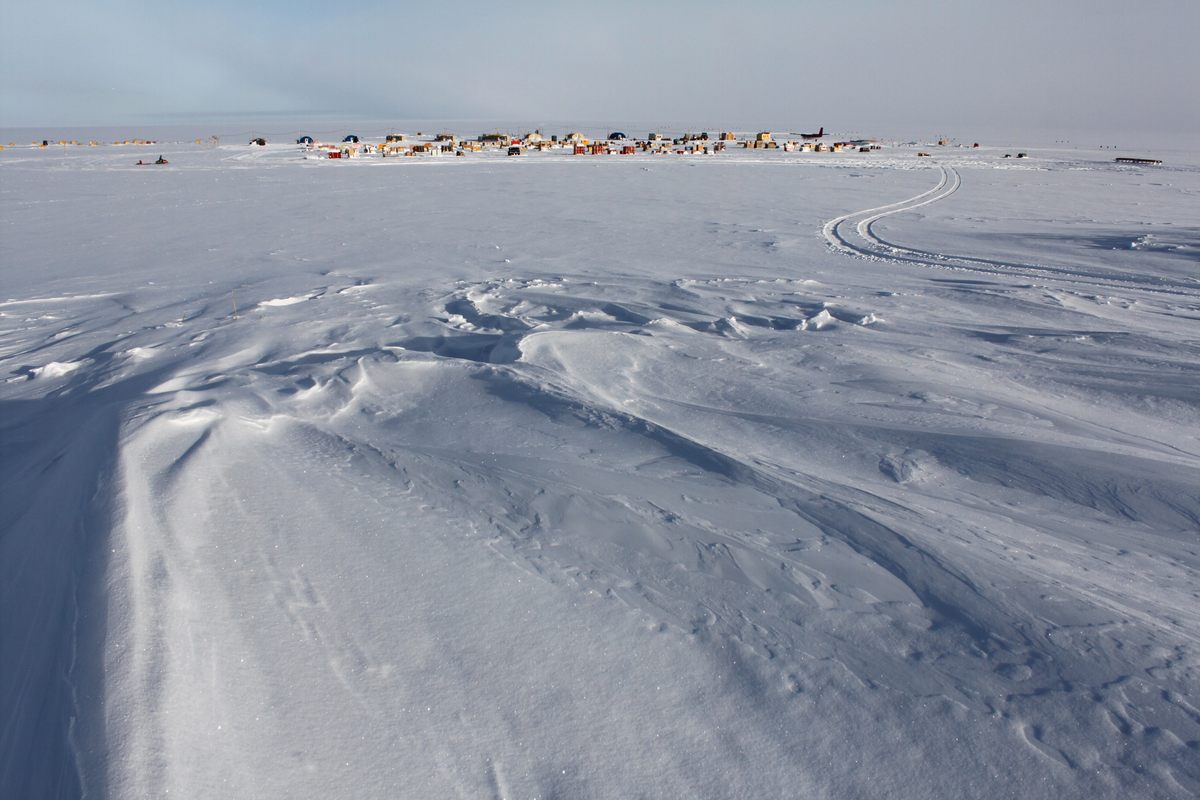 A snow-covered landscape with wind-sculpted snow patterns in the foreground and a small colorful village in the distance under a clear sky.