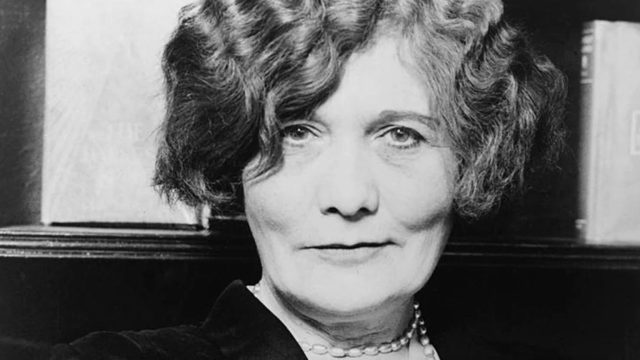 Black and white portrait of a smiling woman with wavy hair and a pearl necklace, circa early 20th century.