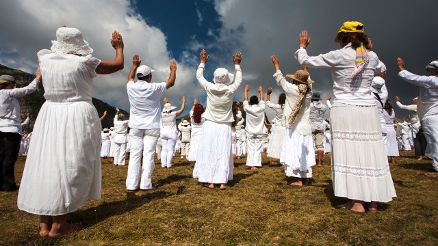 A group of cult members dressed in white participating in an outdoor gathering or ceremony under a partly cloudy sky.