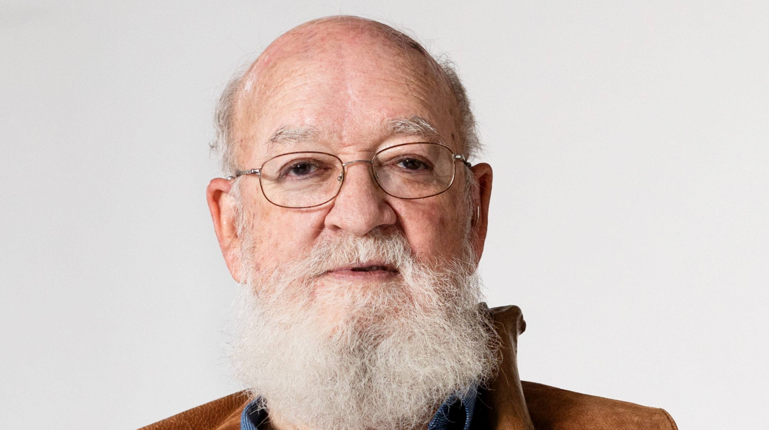 Elderly man with a full white beard and glasses, wearing a brown jacket, against a white background.