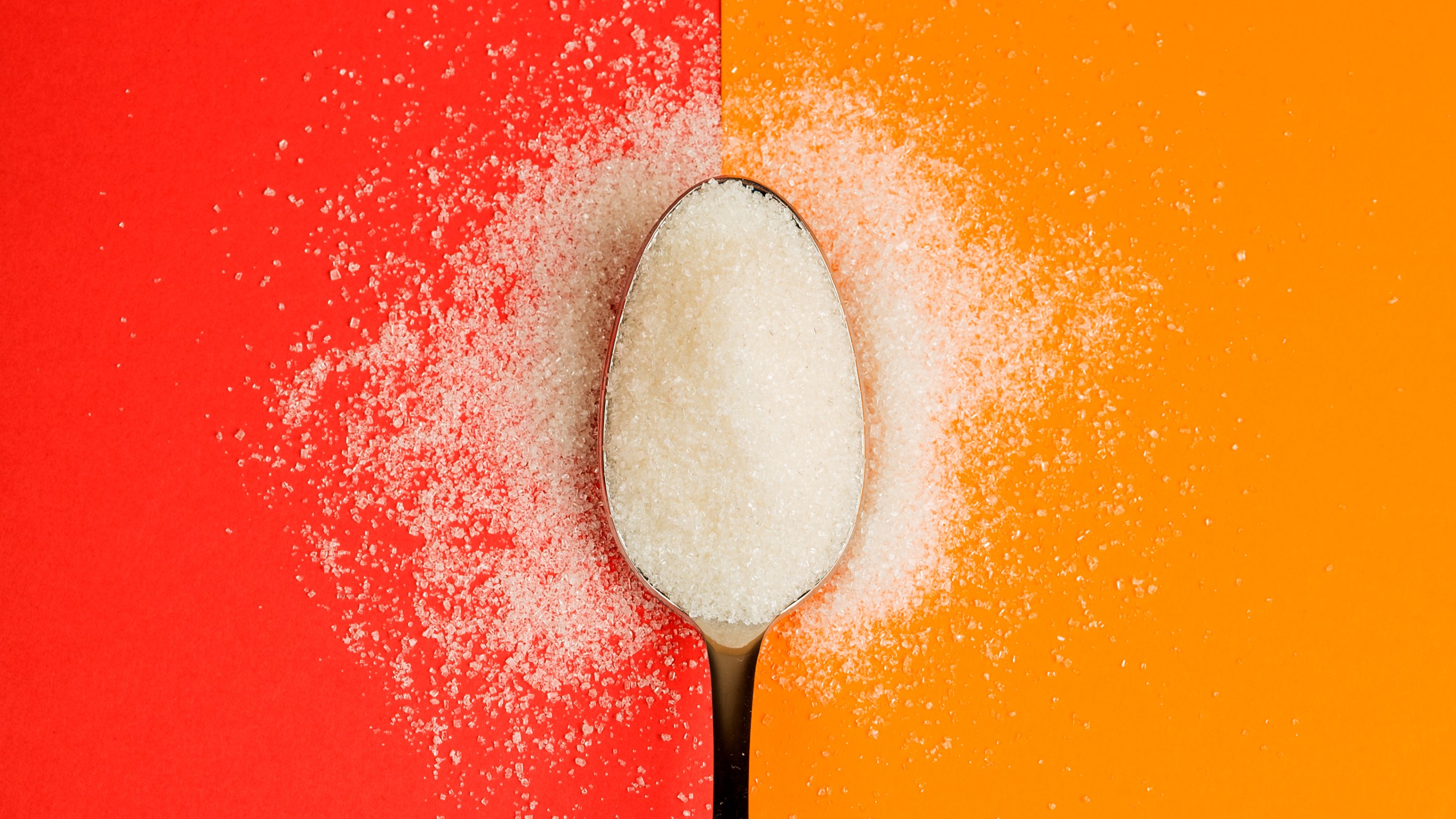 A spoonful of non-sugar sweeteners on a dual red and orange background, with sweetener grains scattered around the spoon.