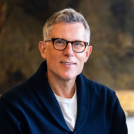 Middle-aged caucasian man with glasses, wearing a navy cardigan and white shirt, smiling and sitting in front of an abstract painting.