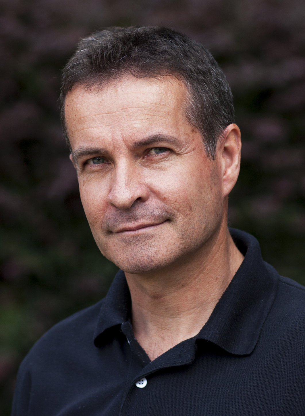 A portrait of a middle-aged man with a slight smile, wearing a black polo shirt, against a blurred natural background.