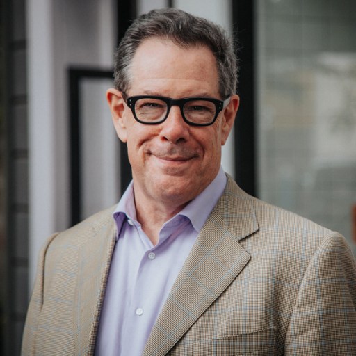 Middle-aged man smiling, wearing glasses, a beige blazer, and a purple shirt, standing outside a building.