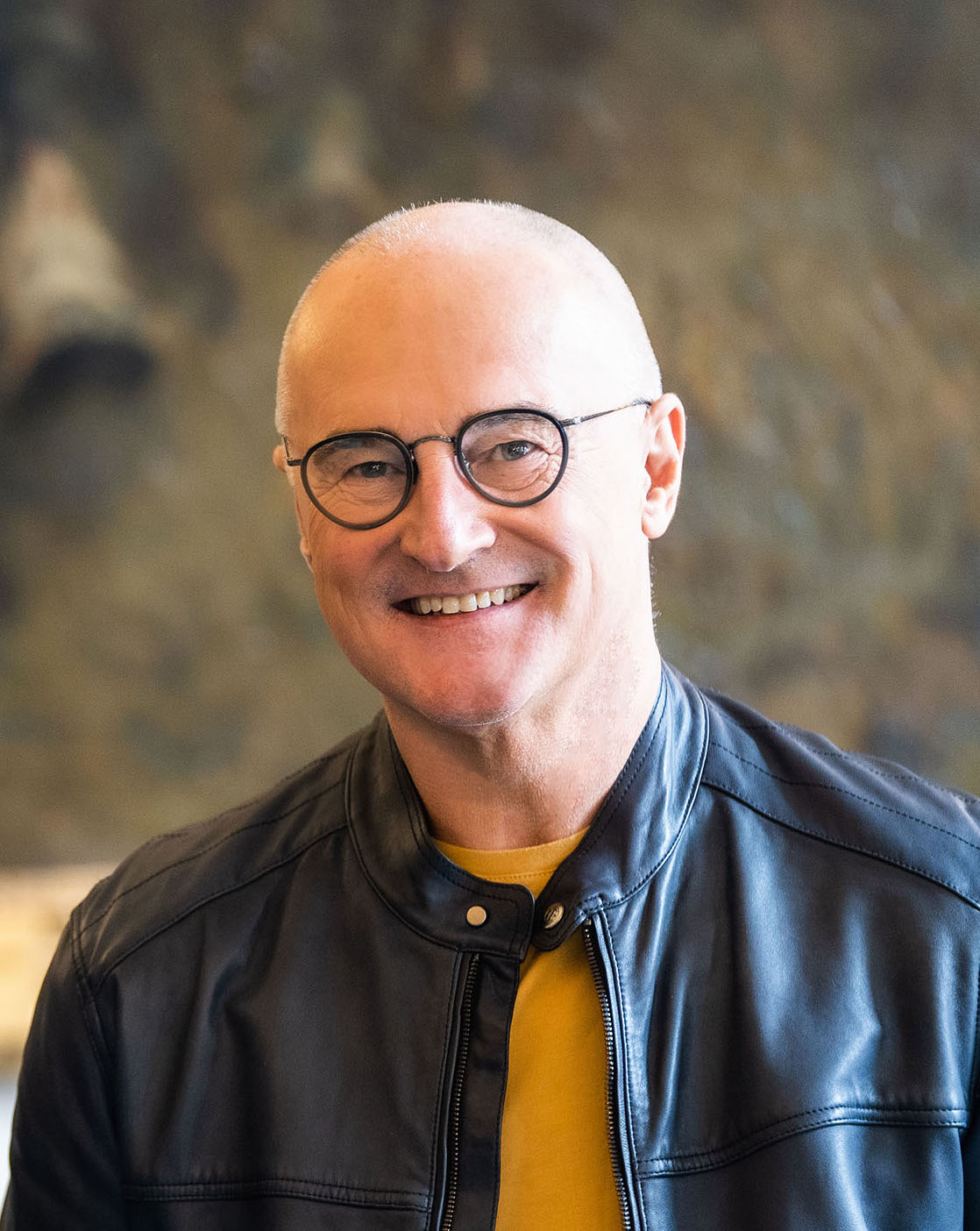 A smiling bald man with glasses, wearing a black leather jacket over a yellow sweater, in front of a blurred background.