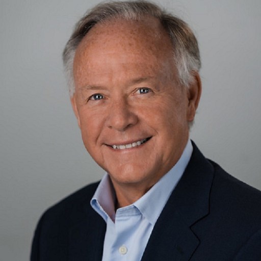 Professional headshot of an elderly man with fair skin smiling, wearing a dark suit jacket and a light blue shirt against a gray background.