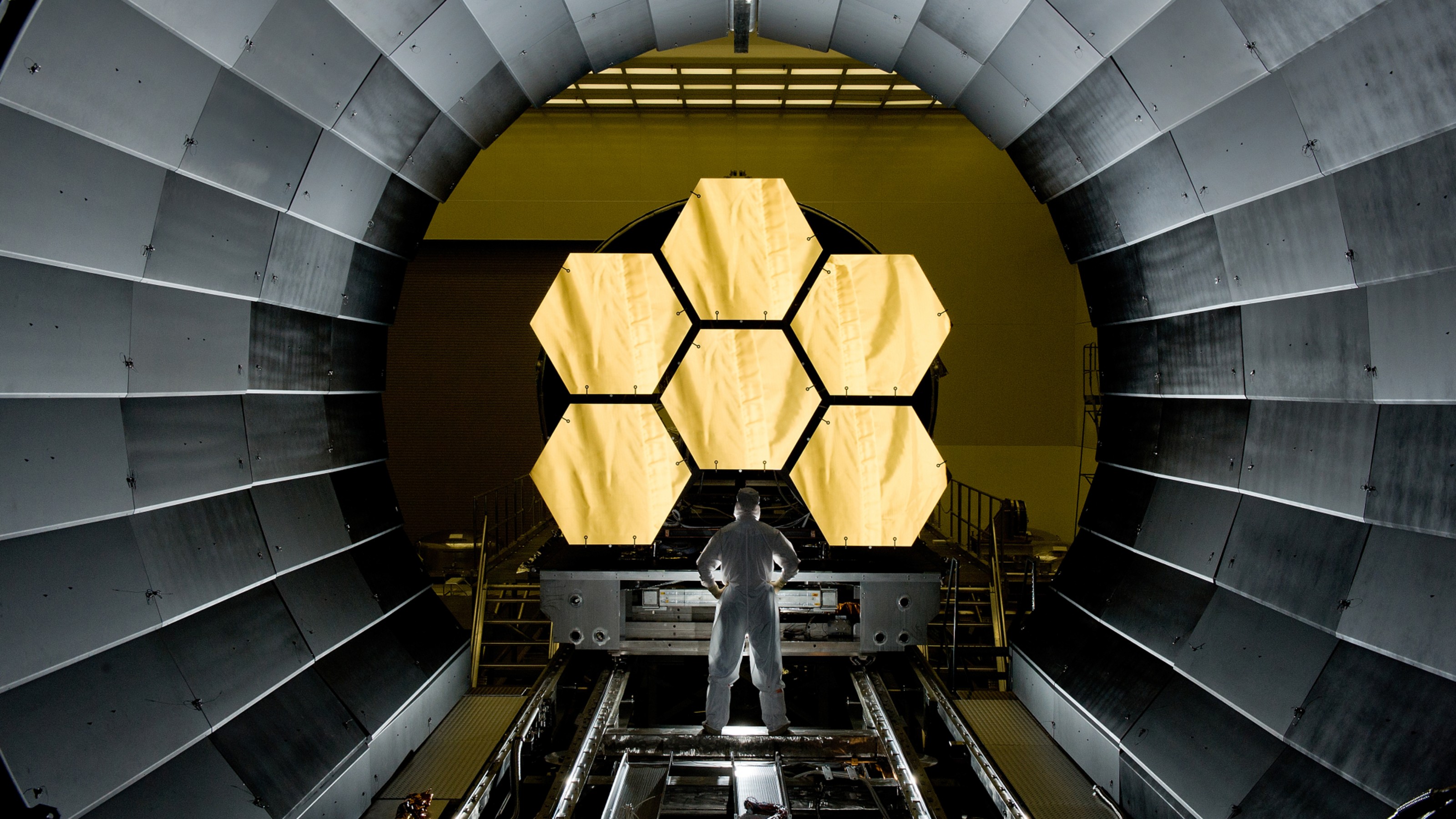 A technician in a clean suit inspects a large, segmented, hexagonal mirror inside a circular gray structure.