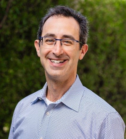 A smiling man with glasses wearing a blue patterned shirt, standing outdoors with green foliage in the background.