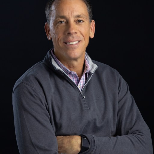 A middle-aged man smiling, wearing a grey pullover and a checkered shirt, arms crossed, against a black background.