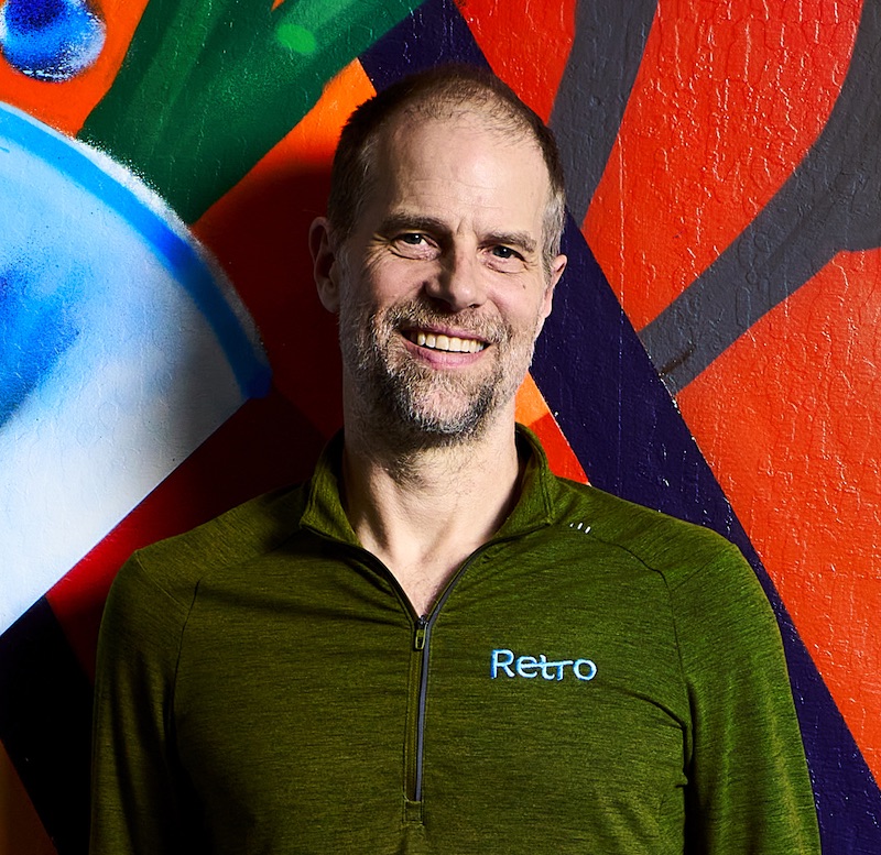 A smiling man with a beard in a green zip-up top stands before a colorful graffiti wall.
