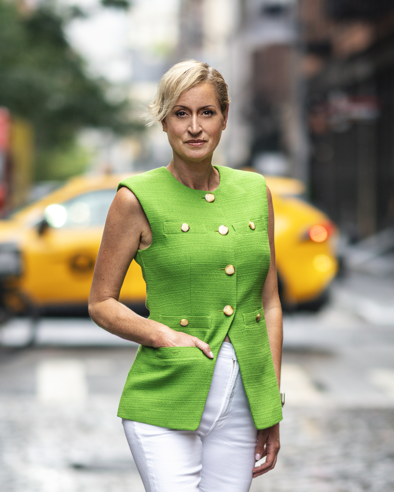 A woman in a green sleeveless top and white pants standing on a city street with a taxi in the background.