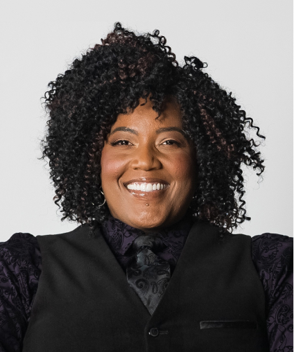 Portrait of a smiling woman with curly black hair, wearing a black ruffled blouse and blazer, against a white background.