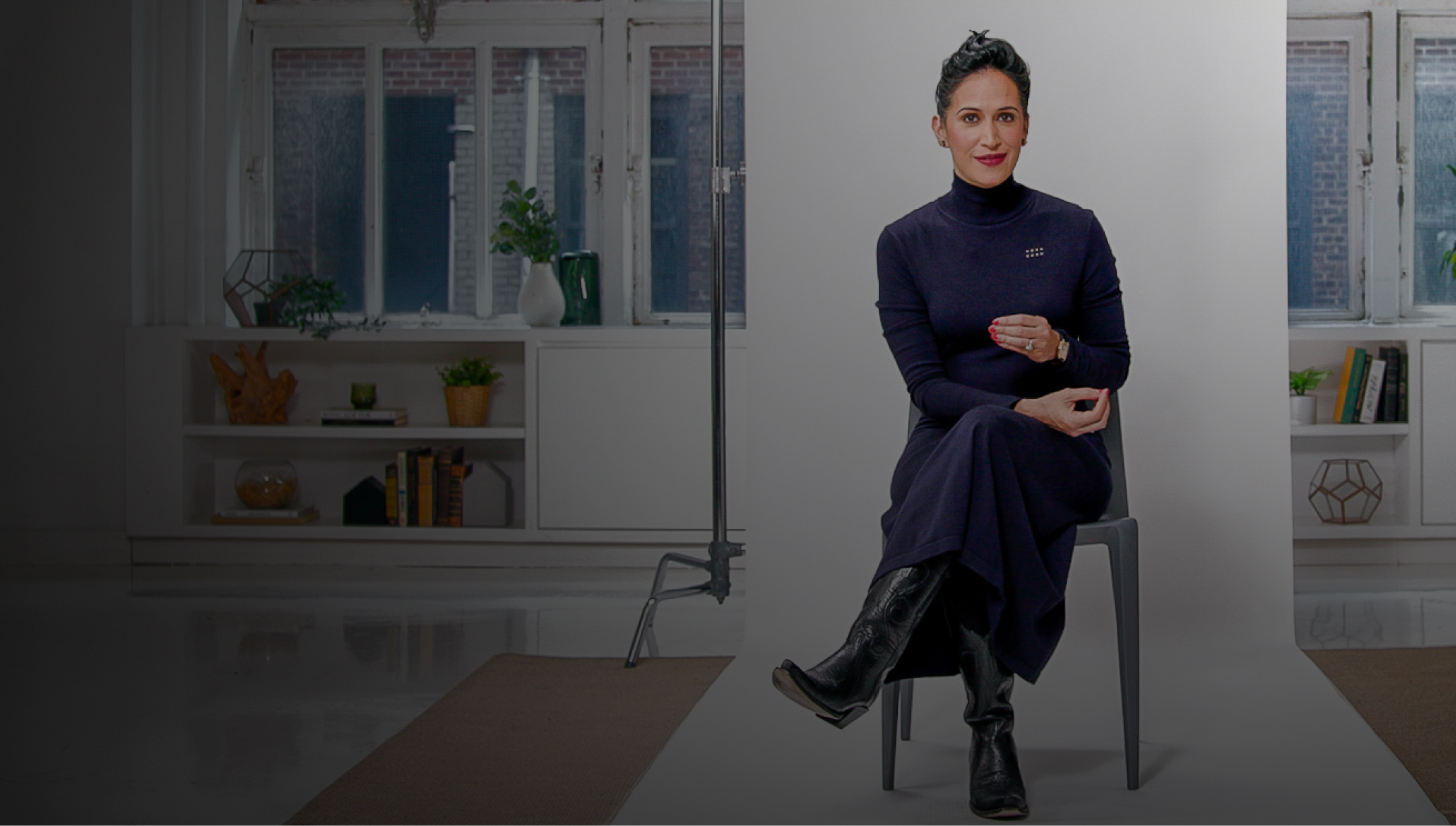 A woman in a dark turtleneck and long skirt sits on a stool in a stylish room with bookshelves, holding a cup, looking at the camera.