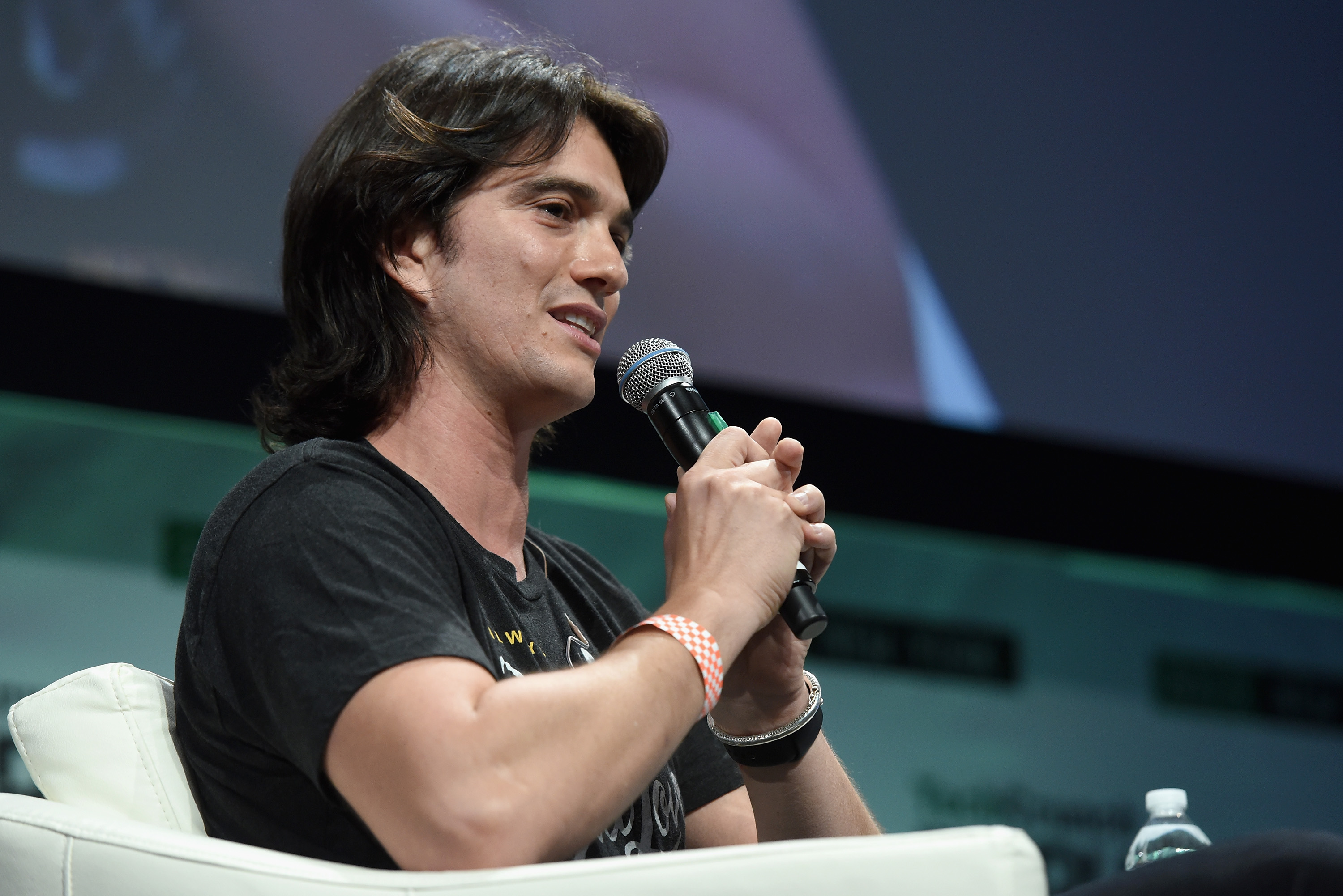 A man with dark hair speaking into a microphone while seated on stage, wearing a black t-shirt and holding a water bottle.