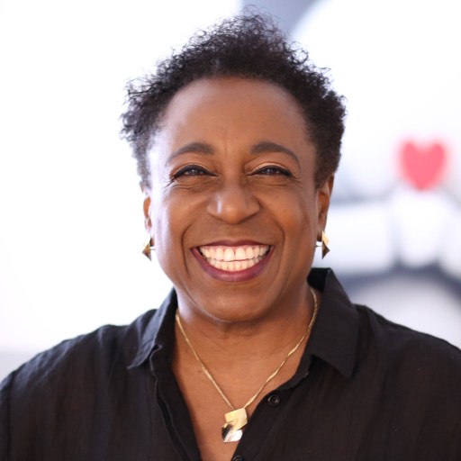 A joyful middle-aged woman with short curly hair, wearing a black shirt and earrings, smiles warmly at the camera.