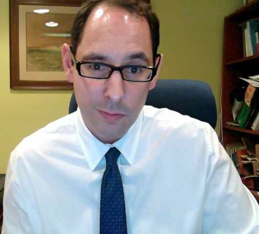 A man with glasses wearing a white shirt and tie, seated in an office with books and a framed painting in the background.