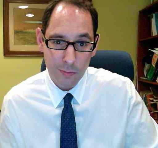 A man with glasses wearing a white shirt and tie, seated in an office with books and a framed painting in the background.