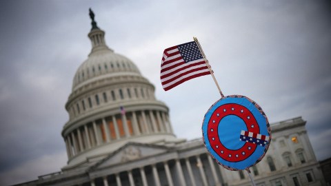 An American flag and a decorative shield with a peace symbol, evoking a sense of paranoia, in front of the United States Capitol building under a cloudy sky.