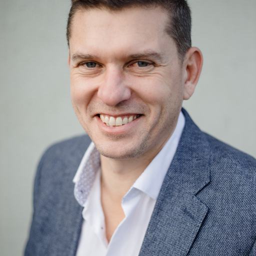 A smiling man in a blue blazer and white shirt against a neutral background.