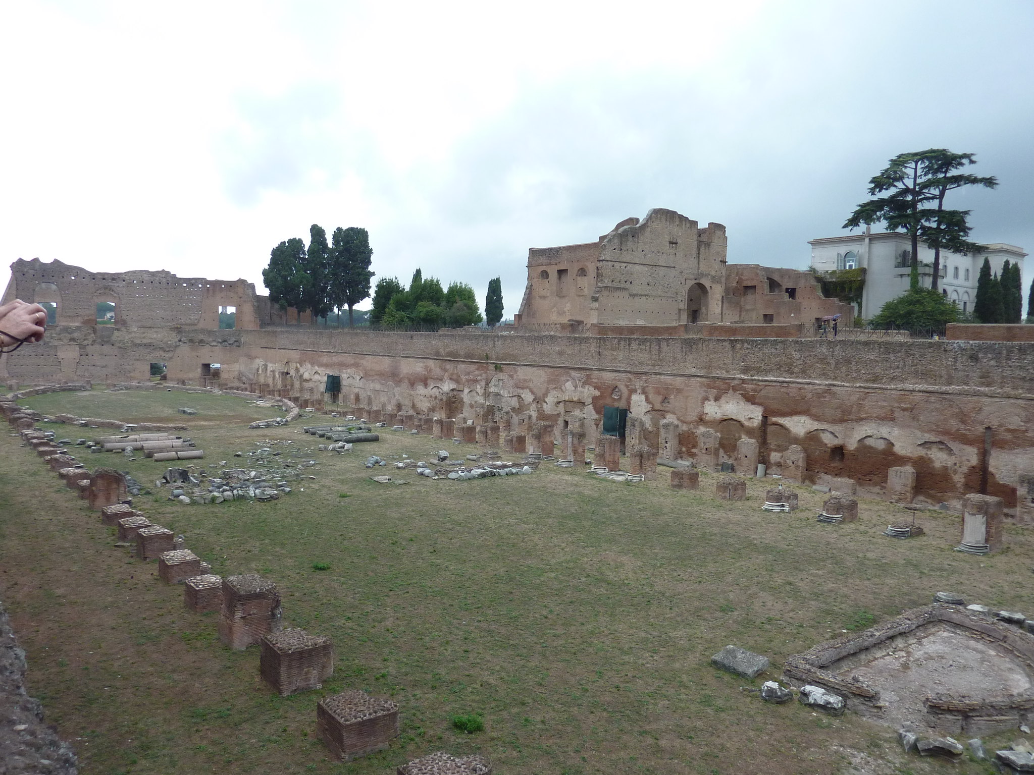 View of ancient Roman ruins featuring large stone structures, a grassy area with scattered stone remnants, and a cloudy sky in the background. A person's hand is visible on the left edge of the image.