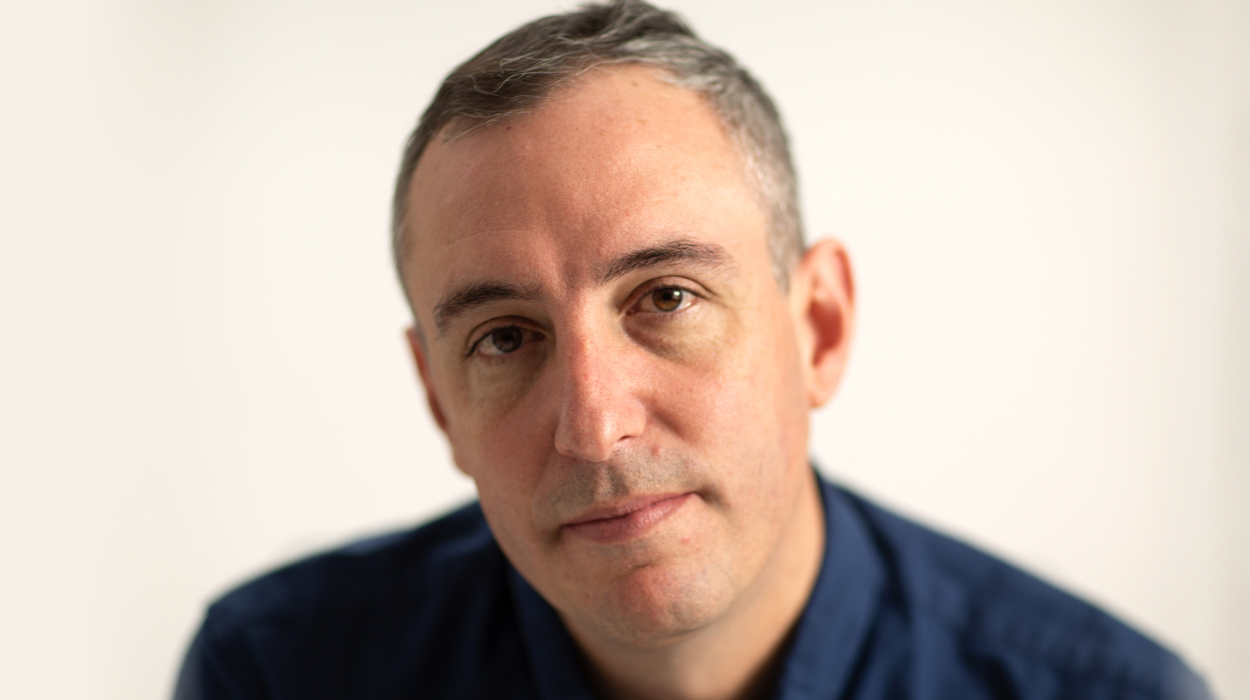 A man with short gray hair and a serious expression is looking directly at the camera, wearing a blue shirt against a plain background.