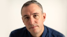 A man with short gray hair and a serious expression is looking directly at the camera, wearing a blue shirt against a plain background.