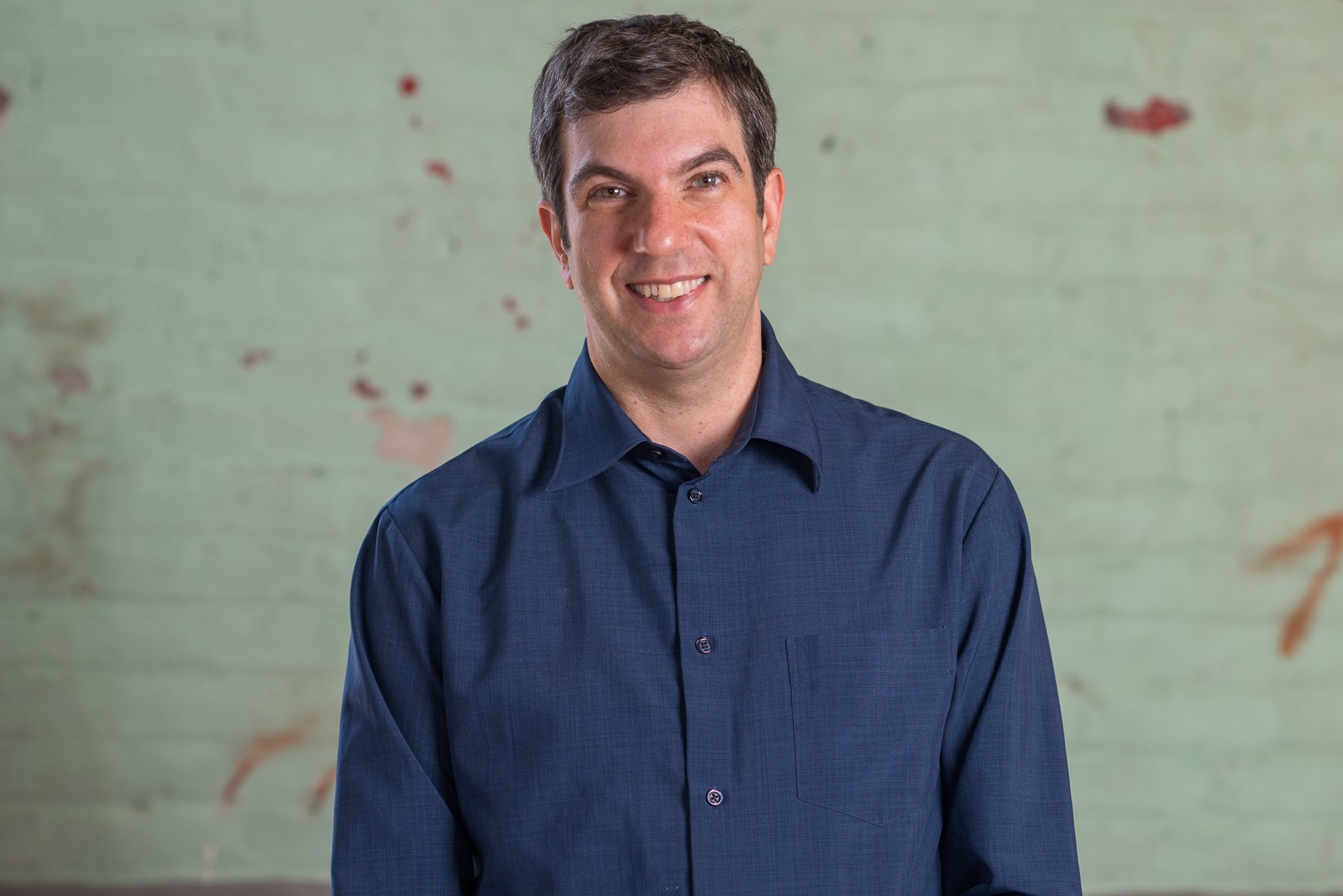 A man in a dark blue shirt smiling at the camera, standing in front of a weathered green wall with red splotches.