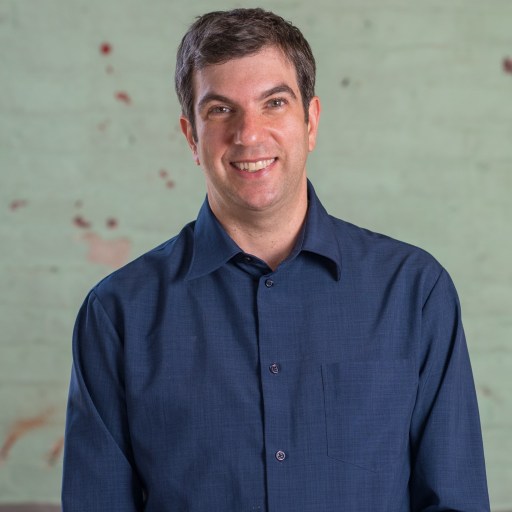 A man in a dark blue shirt smiling at the camera, standing in front of a weathered green wall with red splotches.