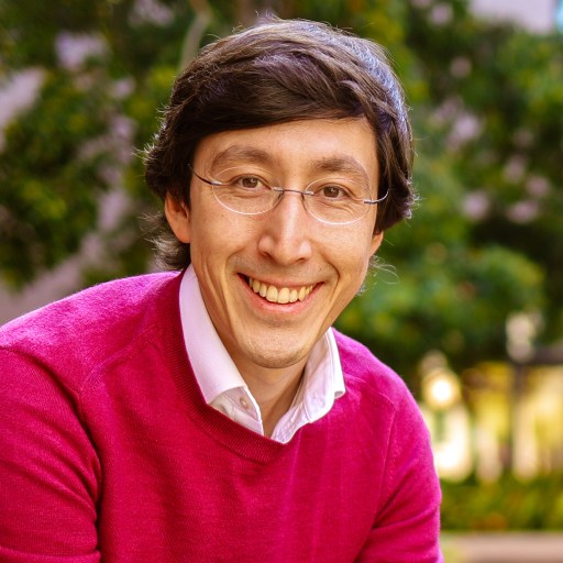 A person with glasses and dark hair, wearing a pink sweater over a white shirt, smiles while sitting outdoors with green foliage in the background.