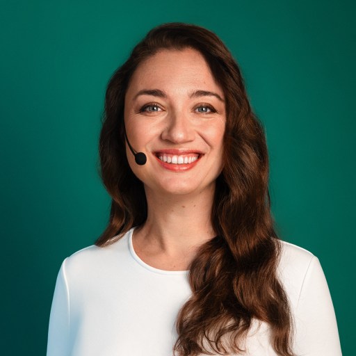 A woman with long brown hair, wearing a white top and a headset, smiles at the camera against a green background.