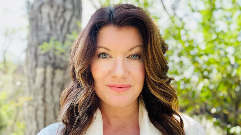 A woman with long, wavy brown hair wearing a white top stands outside in a forested area with green foliage and a tree trunk visible in the background.