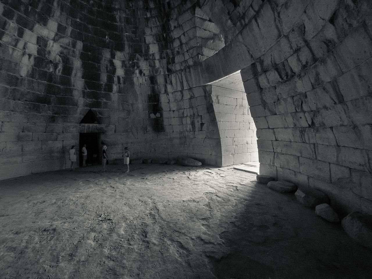 A group of people stand inside a large, ancient stone structure with curved walls and a triangular entrance, illuminated by sunlight from an arched doorway.