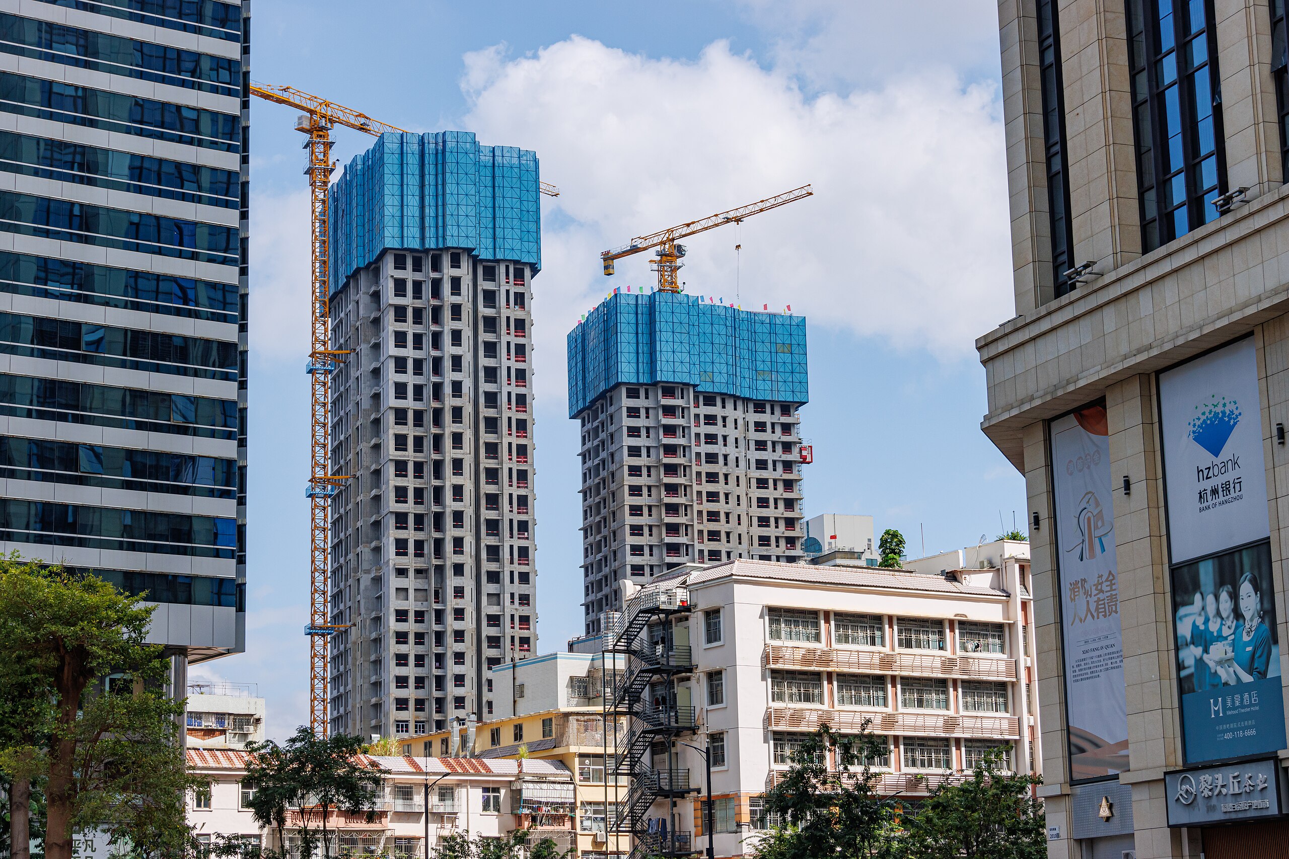 Two partially constructed skyscrapers wrapped in blue protective netting, surrounded by buildings, cranes, and trees, under a blue sky with clouds.