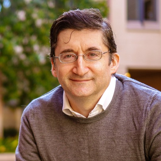 A man with glasses, short dark hair, and a gray sweater over a white collared shirt smiles outdoors in front of a blurred background of greenery and a building.