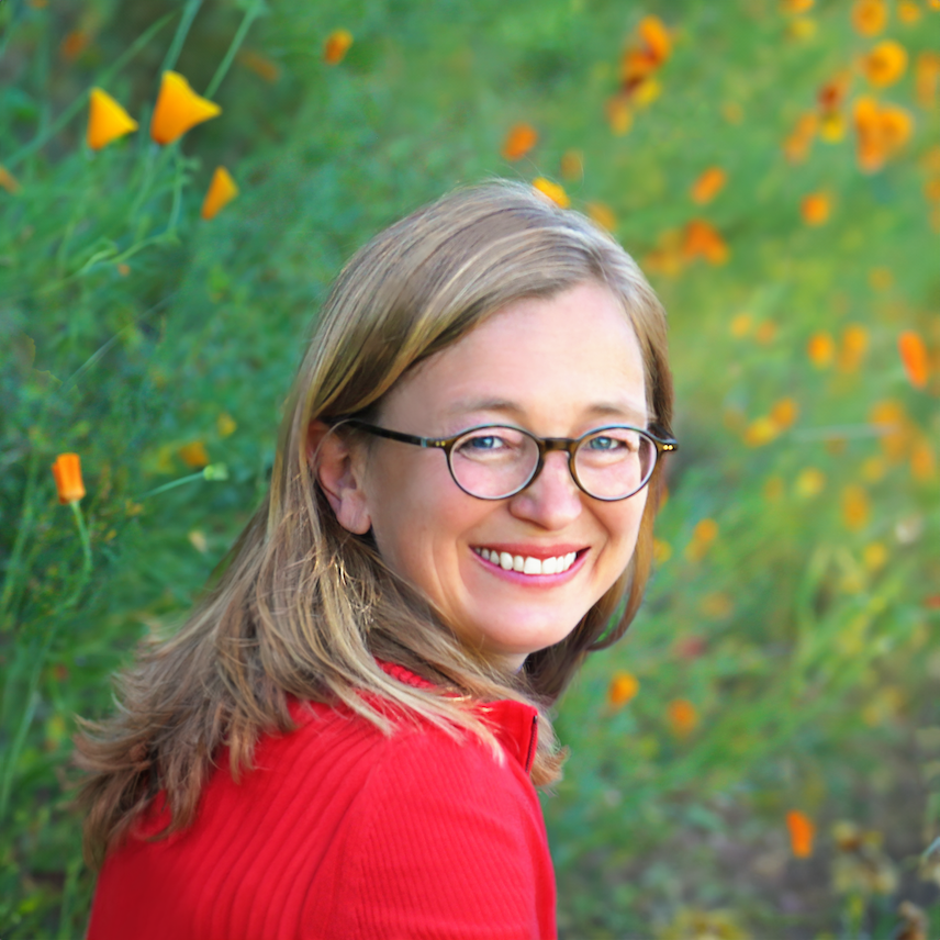 A woman with glasses and a red sweater is smiling while sitting in a field with blooming orange flowers.