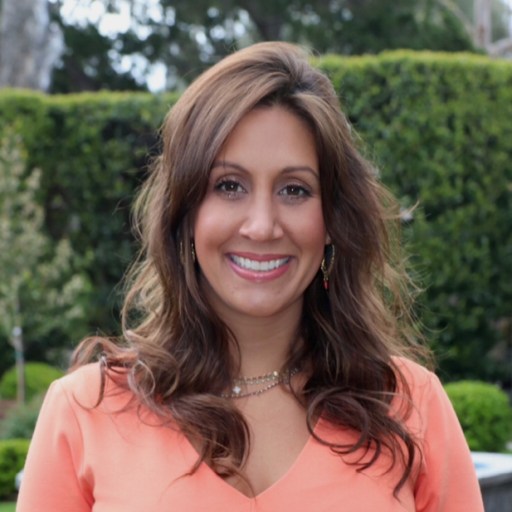 A woman with long brown hair smiling, wearing an orange top and standing outdoors in front of green foliage.