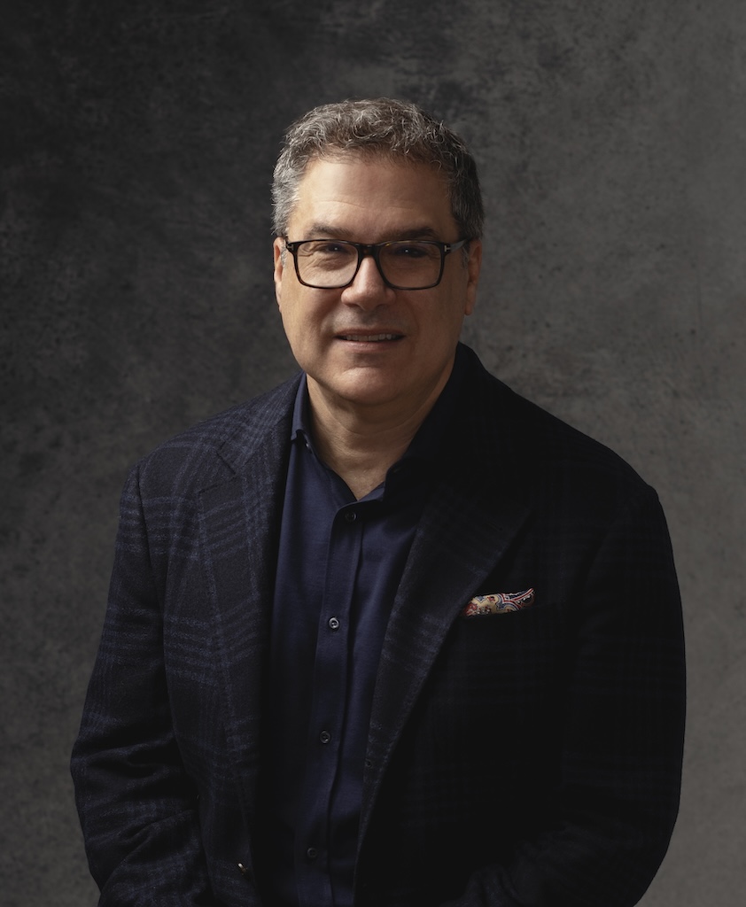 Middle-aged man wearing glasses and a dark blazer, poses against a textured dark gray background.