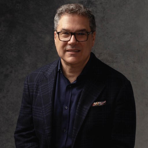 Middle-aged man wearing glasses and a dark blazer, poses against a textured dark gray background.