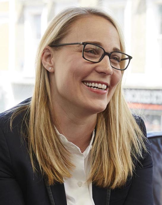 A smiling woman with blonde hair and glasses, wearing a navy blazer and white shirt, seated indoors.